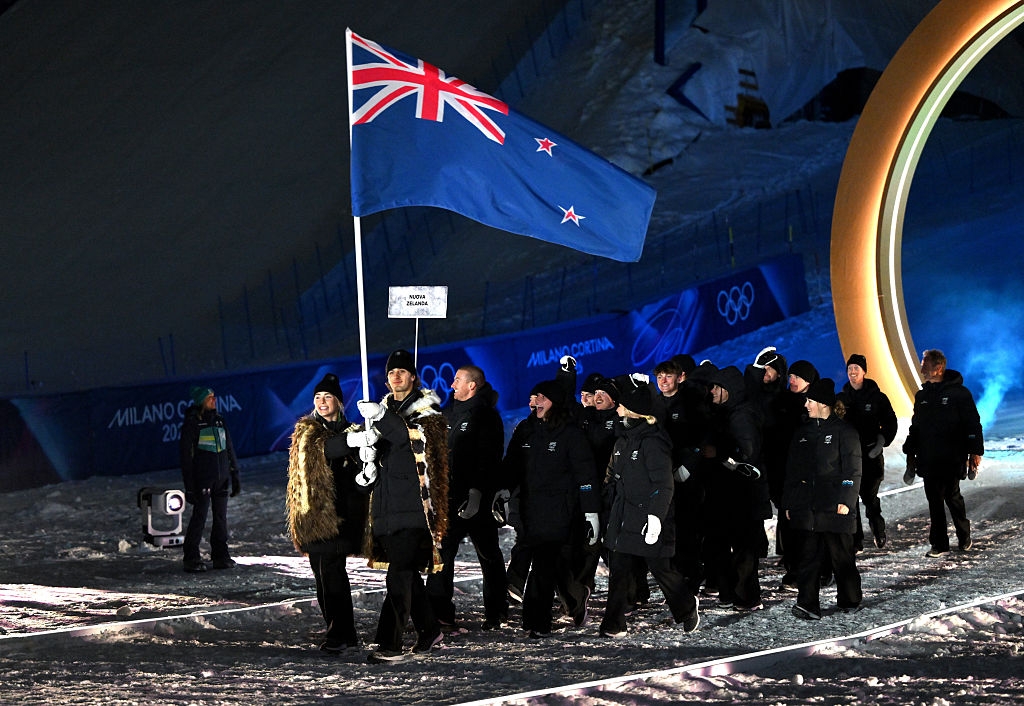 Athletes in winter gear carry a New Zealand flag during an Olympic parade on a snowy path, with a large ring decoration in the background