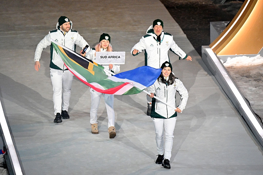 Four athletes in winter sports uniforms carry the South African flag at a ceremony. They're holding a sign that says "Sud Africa."