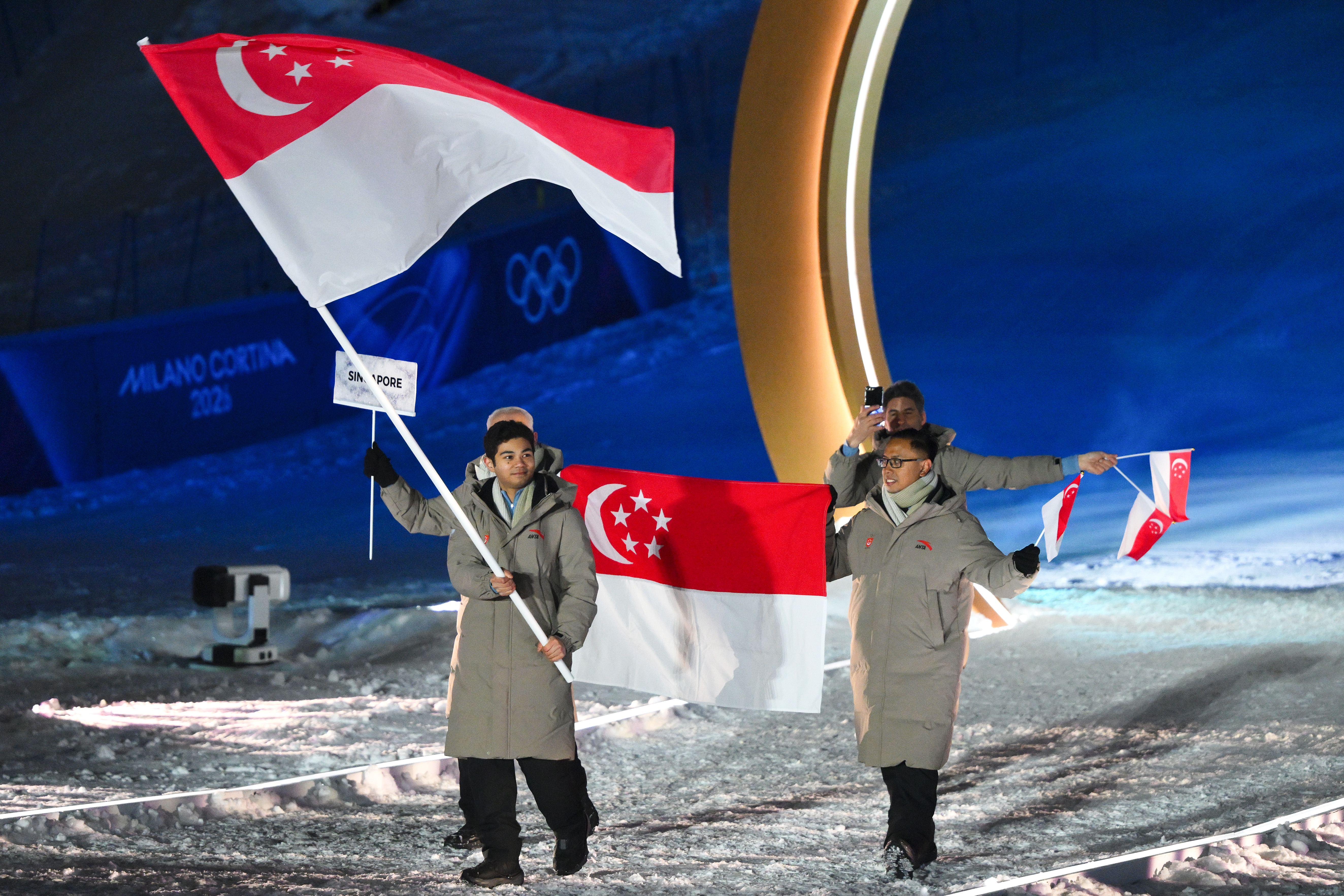 People at the Olympics opening ceremony carry Singapore flags, wearing winter coats. Iconic Olympic rings are visible in the background