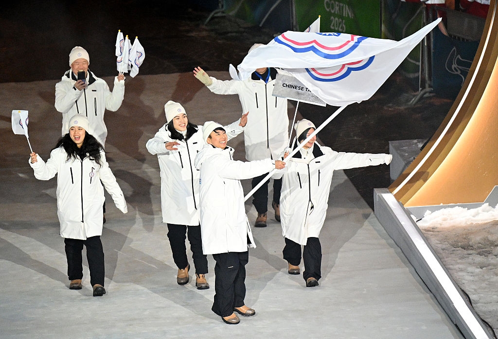Group of athletes in white winter attire waving flags at a sporting event, leading a parade-like entrance