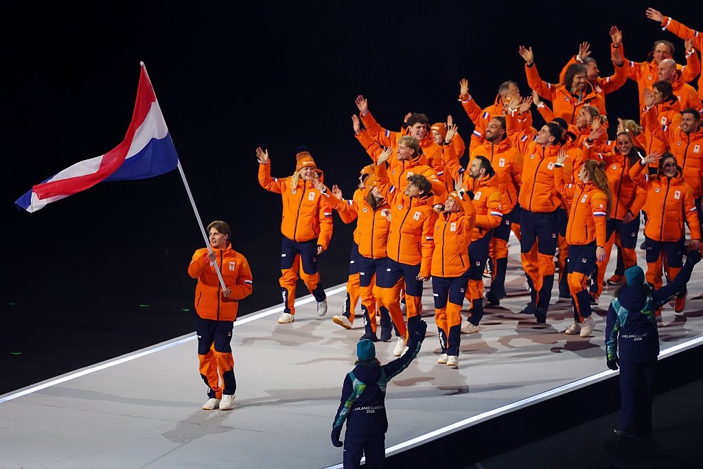People in matching winter outfits walk onstage, waving. One person holds a flag with horizontal stripes