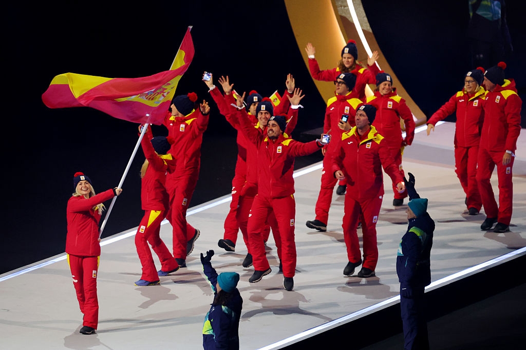 Group of athletes in matching outfits, waving and holding a flag, march in a sports event arena, with others cheering nearby