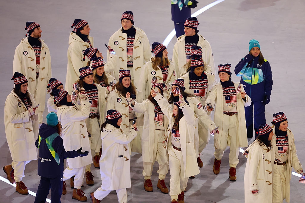 Athletes in matching winter outfits with American flags, wearing boots and beanies, walk together in a stadium setting