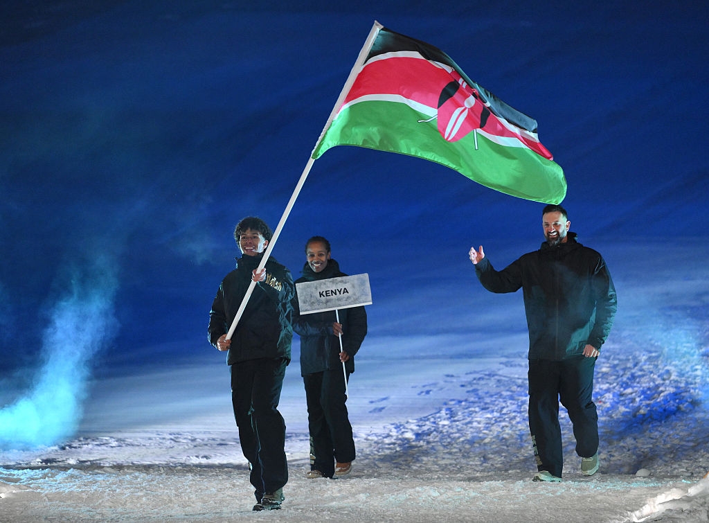 Three individuals walk on a snowy path, with one holding the Kenyan flag and another holding a "Kenya" sign. They wear winter attire