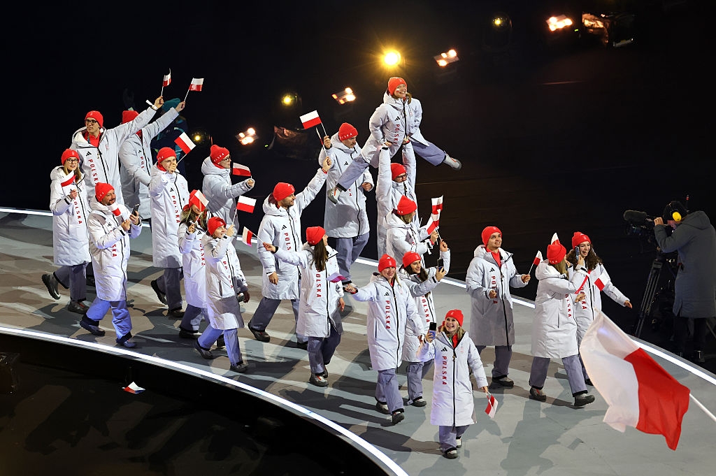 Athletes march in a stadium wearing winter jackets and red hats, waving flags, during an opening ceremony parade