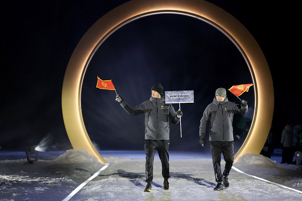 Two people in winter outfits walk through a circular archway, holding Montenegro flags and a sign during an outdoor ceremony or event