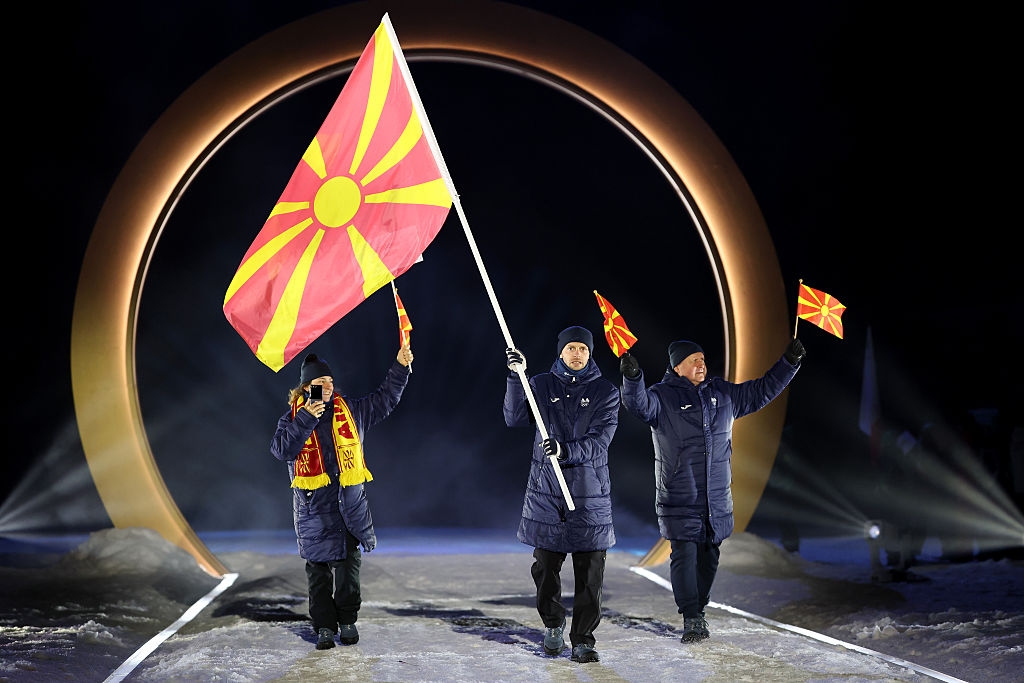 Three people in winter clothing carry flags with a circular backdrop, participating in a formal outdoor event or ceremony