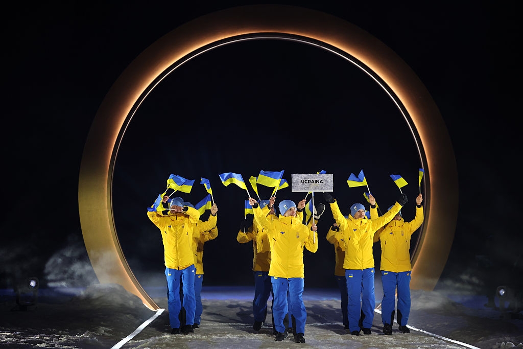 People in coordinated yellow and blue outfits wave flags, entering through a large circular arch with a sign reading "Ukraine."