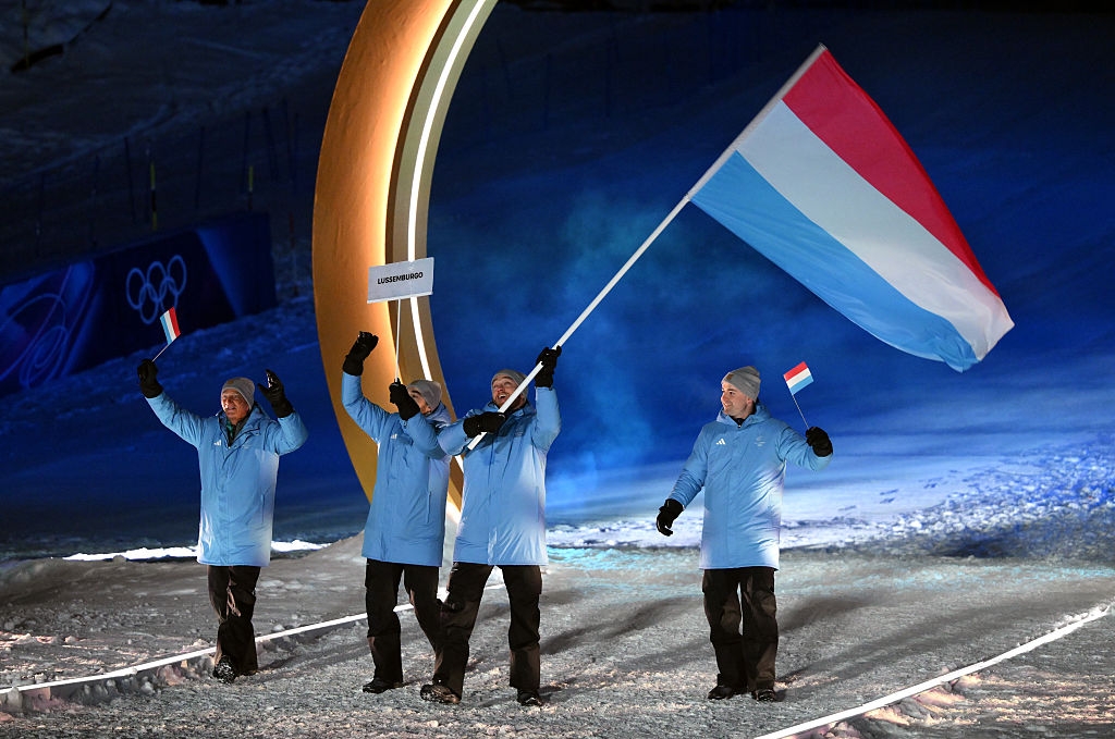 Four athletes in matching uniforms wave flags at a winter sporting event, standing near a lit Olympic ring structure