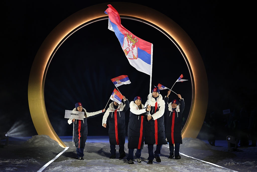 Group of athletes in matching long coats wave Serbia's flags while entering an arena, holding a large national flag prominently