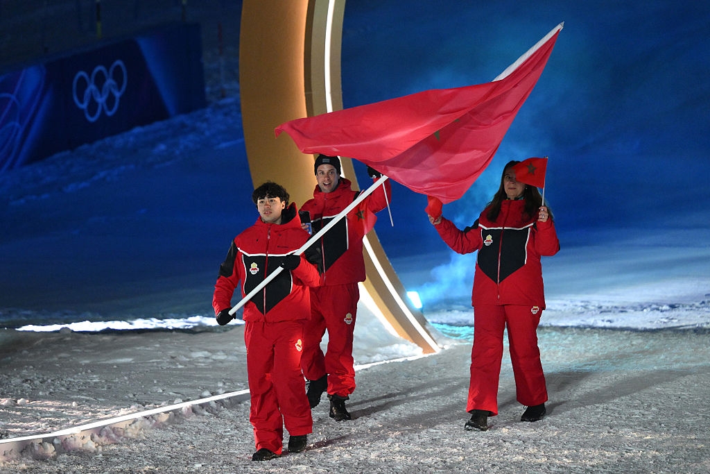 Athletes in red winter attire carry their country's flag during an Olympic ceremony on a snow-covered stage