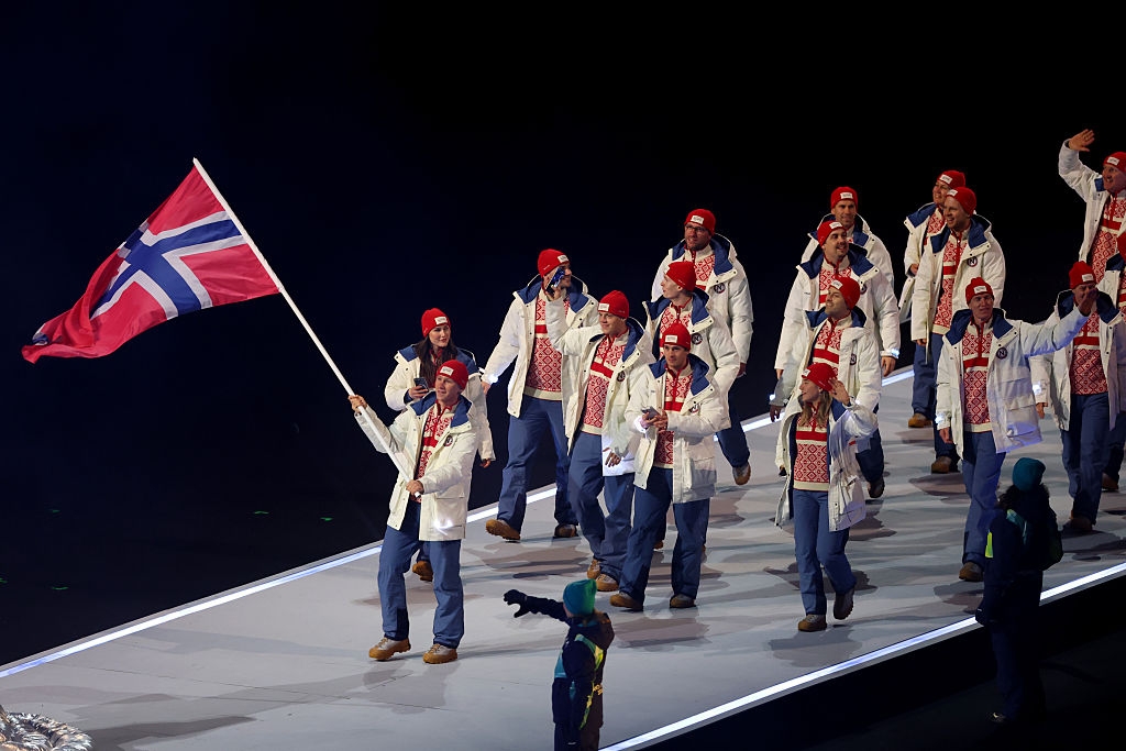 Norwegian athletes in patterned sweaters and jackets walk on stage, one holding the national flag, at a sports event