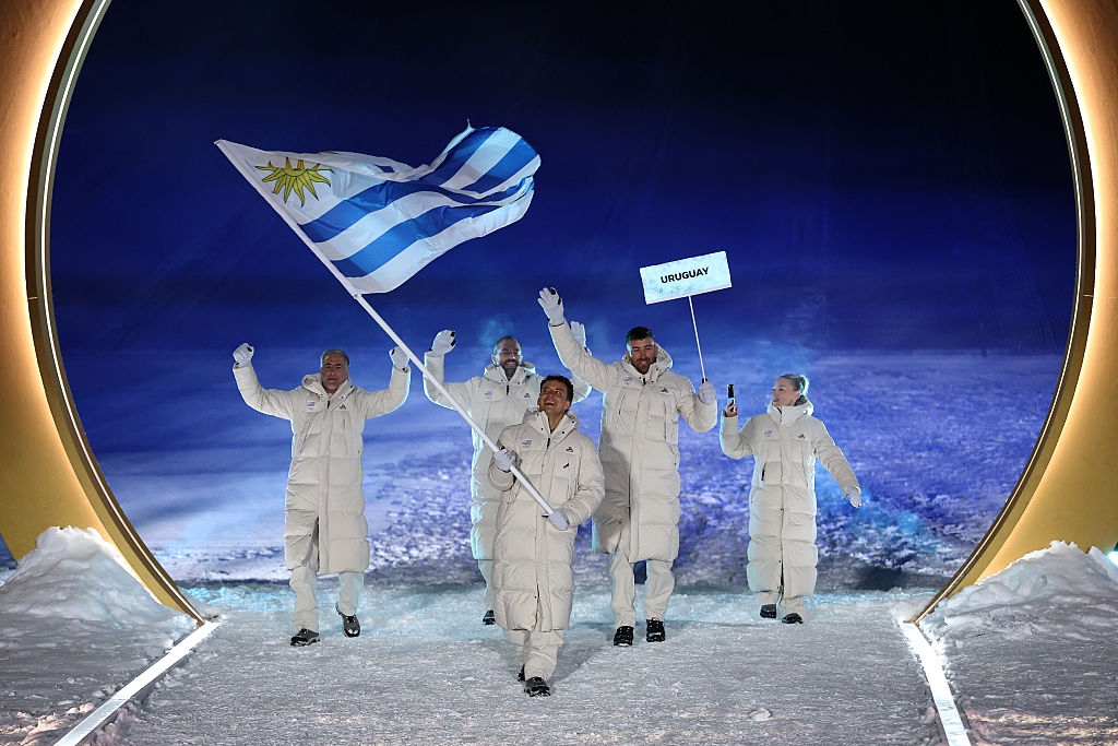People in white winter clothing walk through a snowy area carrying the Uruguayan flag and a sign reading "Uruguay."