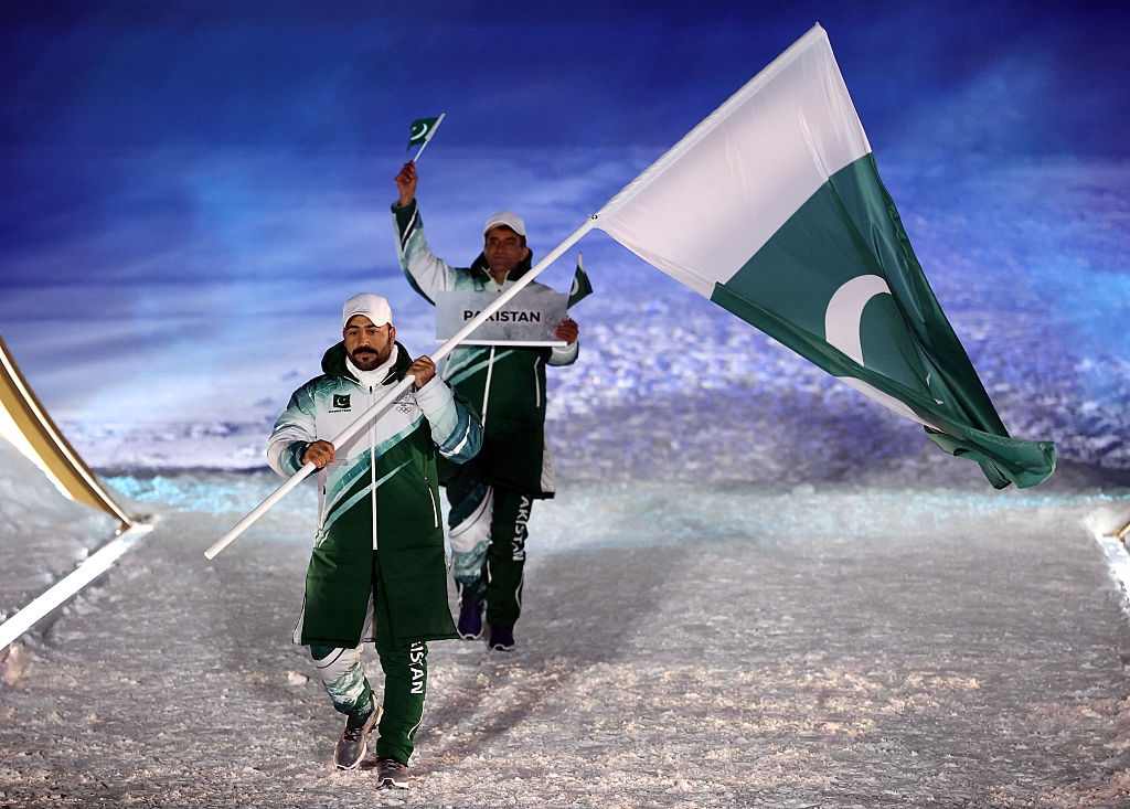 Athletes in matching uniforms walk on a snowy path, each carrying a flag with a large white crescent and star