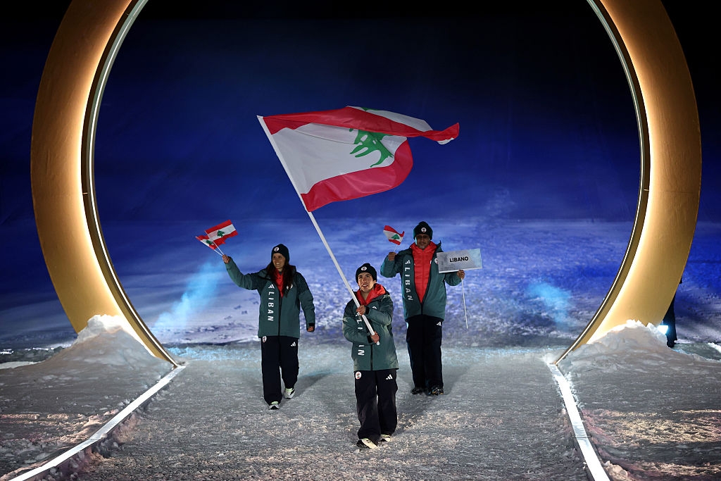 Three individuals, in winter sports attire, carry a large Lebanese flag and a sign reading "Lebanon" at a sports event under a glowing arch