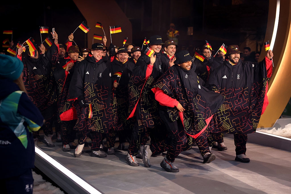 Group of athletes in patterned capes and matching beanies wave flags while walking on a stage during an event
