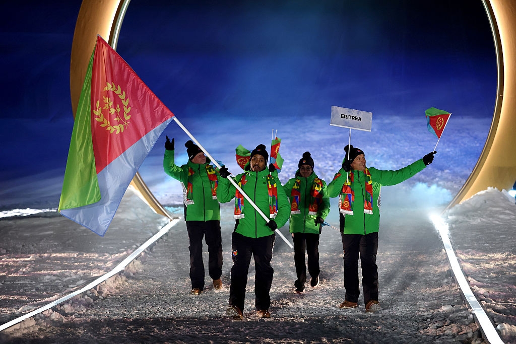 Athletes in green jackets hold Eritrea's flag during an Olympic opening ceremony, marching proudly on a snowy path under a large arch