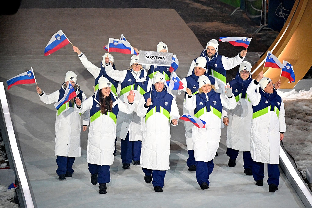 A team of athletes enters an arena during an opening ceremony, wearing matching winter uniforms and waving Slovenian flags