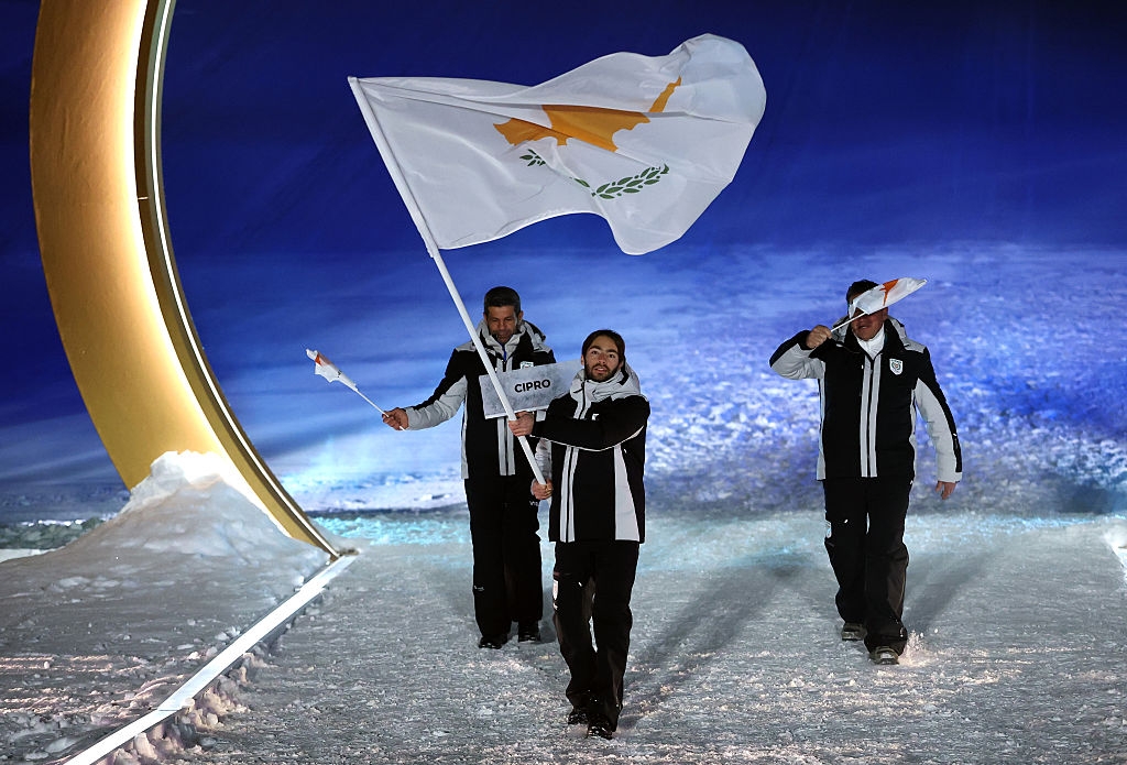 Three athletes in winter gear walk on a snowy path at an event, one holding a Cyprus flag, another saluting, under an illuminated arch