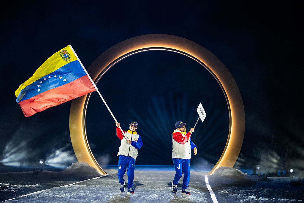 Two individuals in colorful athletic attire walk on a stage holding a Venezuelan flag and a sign, under a large circular arch