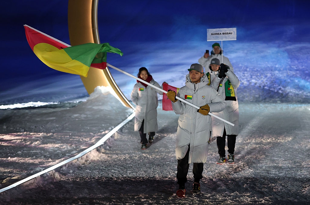 People in white coats walk on snowy ground, one holding a flag. A sign for Guinea-Bissau is visible in the background