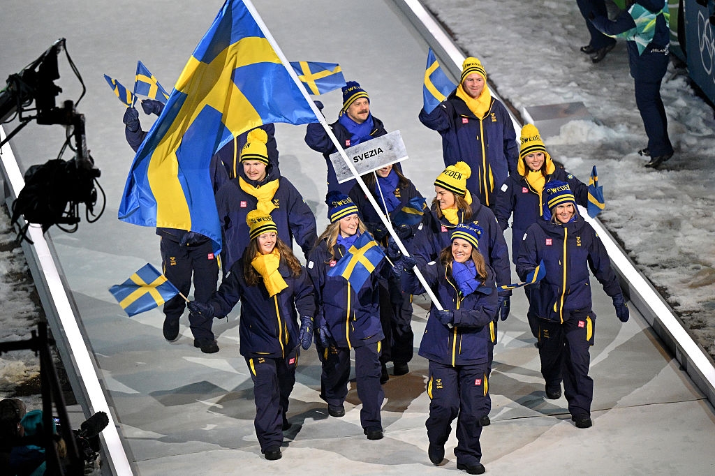 Team Sweden athletes in matching winter uniforms proudly march with flags at an indoor ceremony
