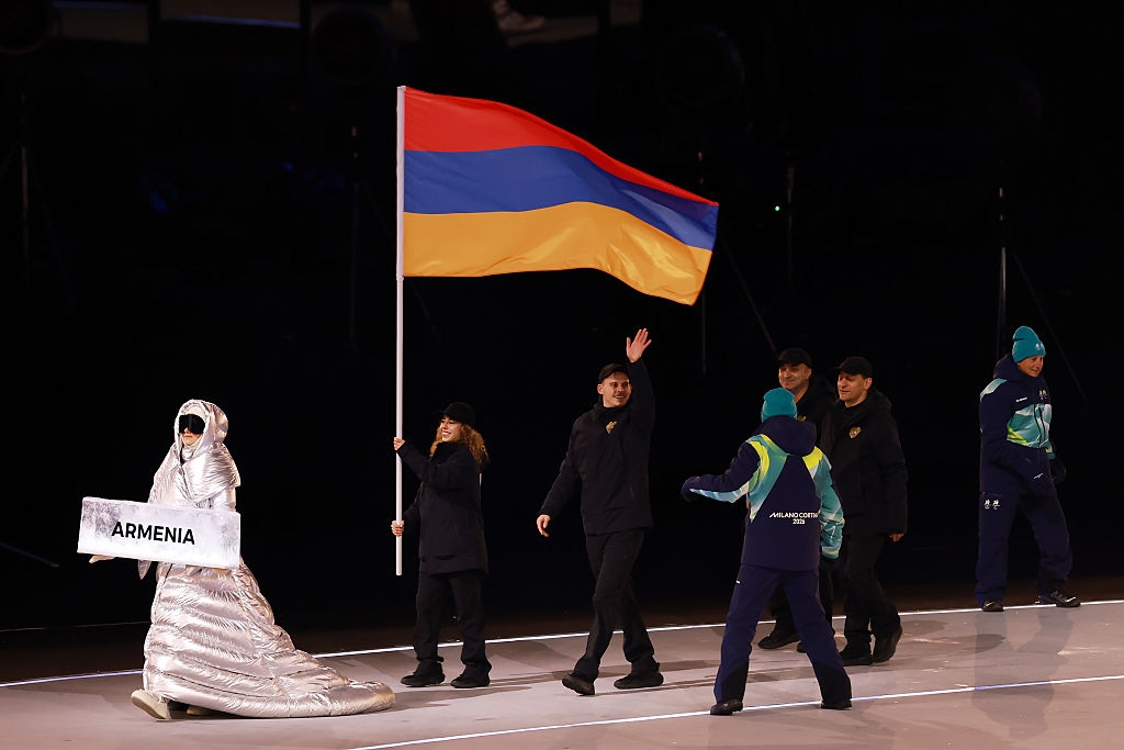 Person waving Armenian flag during a parade, followed by others in dark attire. Another person in shiny outfit holds an "Armenia" sign