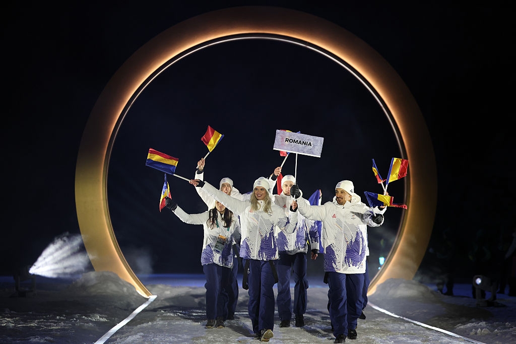 Romanian athletes march during a ceremony, carrying flags. They wear matching jackets and pants, led by a sign bearer in front of an illuminated arch