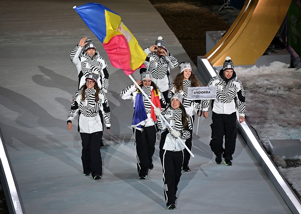 Athletes in zebra-striped outfits march in a parade, waving flags and holding a sign for Andorra