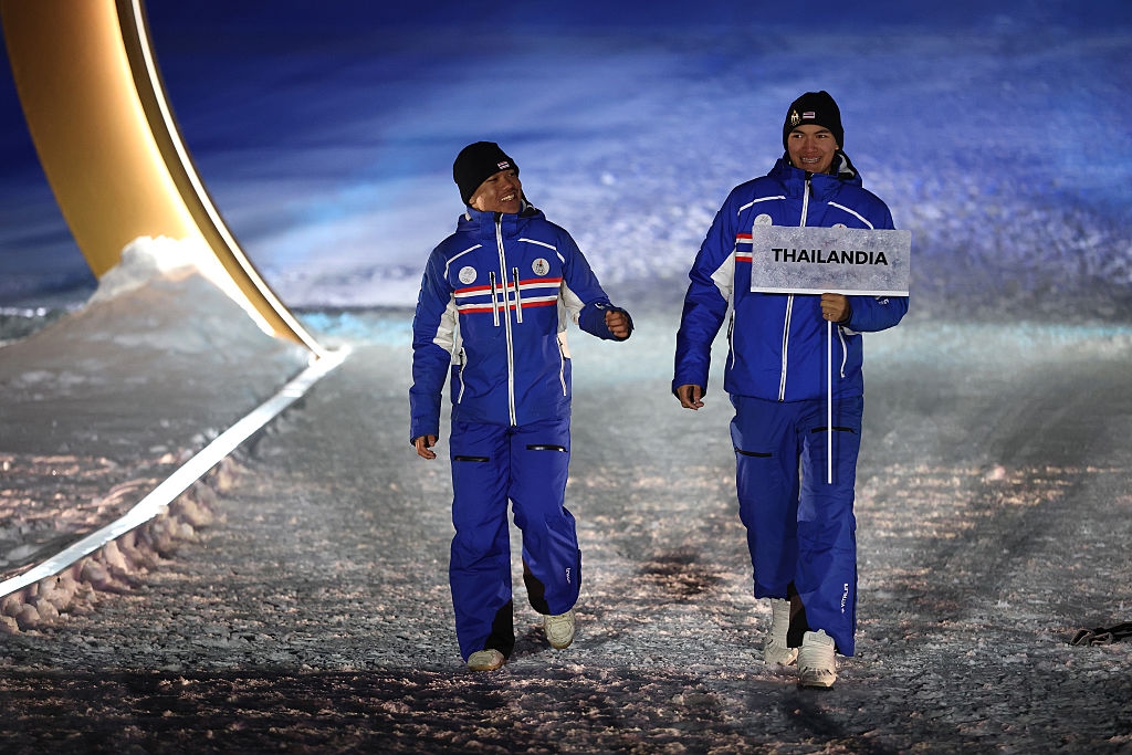 Two athletes in matching winter gear walk on a snow-covered path, one holding a sign that reads "Thailandia."
