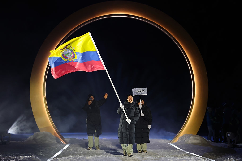 People in coats walk through a large circular structure, one holding the Ecuadorian flag, and another a "Ecuador" sign at a ceremonial event