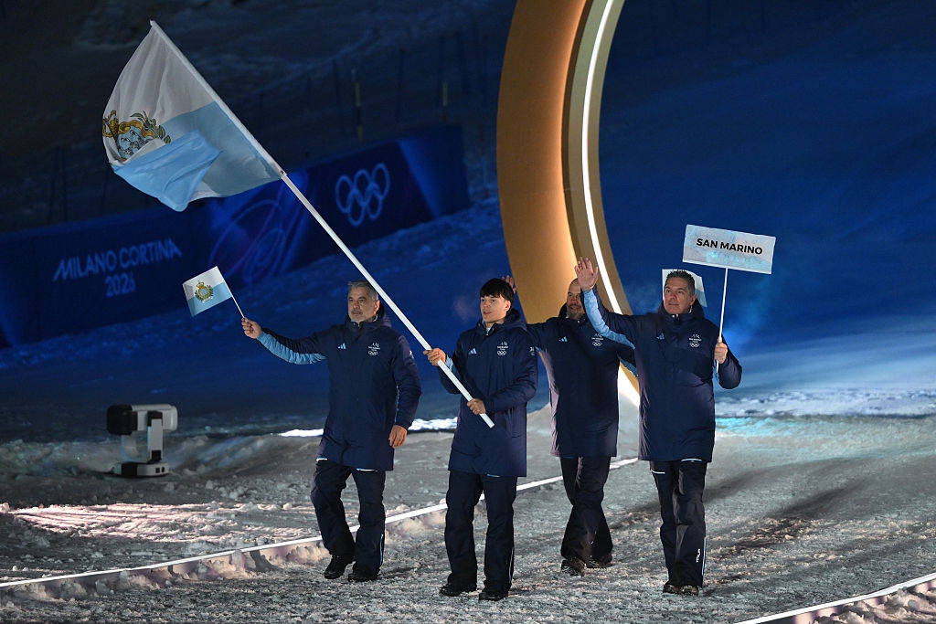 People in matching winter attire hold flags, leading a delegation during the Milan-Cortina Olympics ceremony
