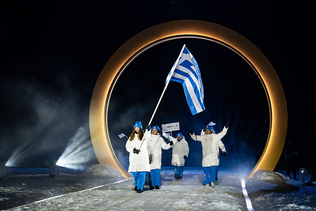 Athletes in white winter gear walk through a lit arch, holding a Greek flag at an Olympic ceremony