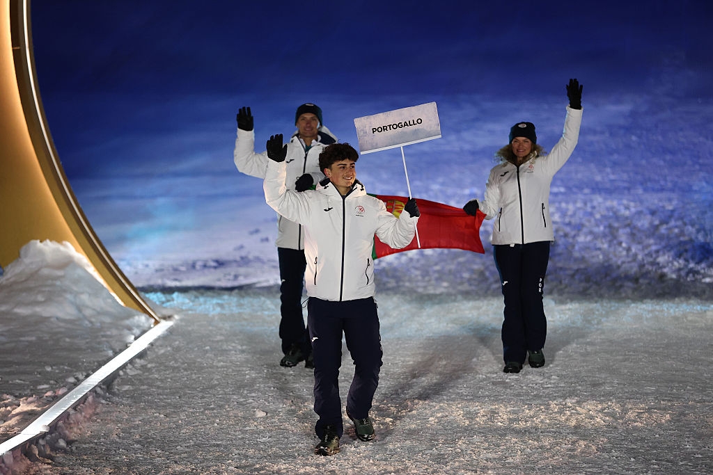 Three athletes in winter attire walk on snow, one holding a "Portogallo" sign, indicating participation in a winter sports event