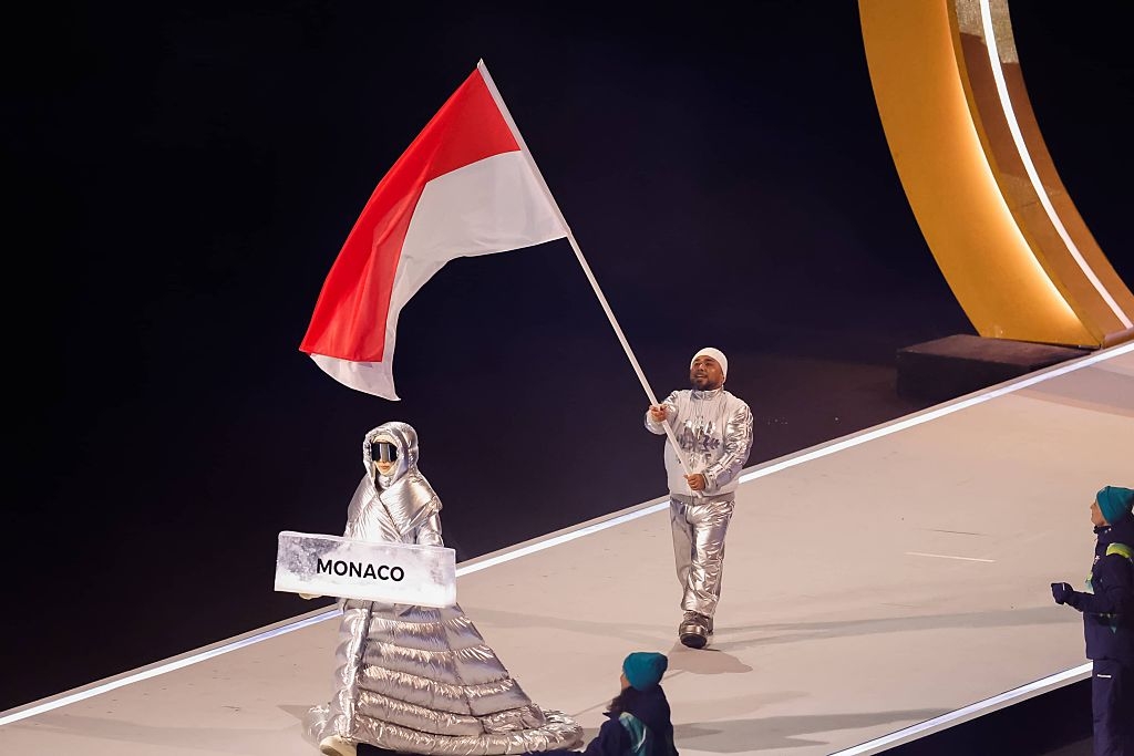 Person in silver quilted gown holds "Monaco" sign. Another in silver suit waves red and white flag. Both walk on a stage, possibly an event or ceremony