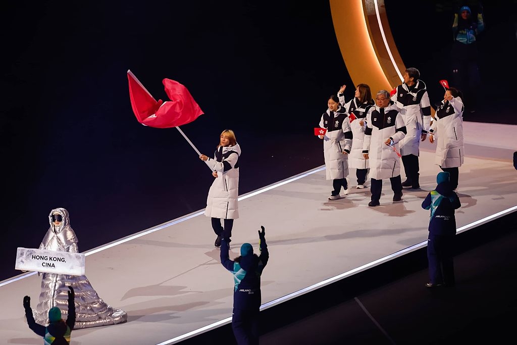 Athletes in white coats hold flags during a parade at a sporting event, with a person leading holding a sign reading "Hong Kong, China."