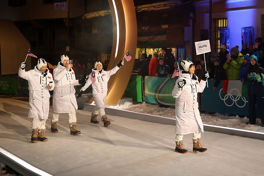 Athletes in white winter gear wave flags during a parade at a snowy sports event, with Olympic rings visible in the background