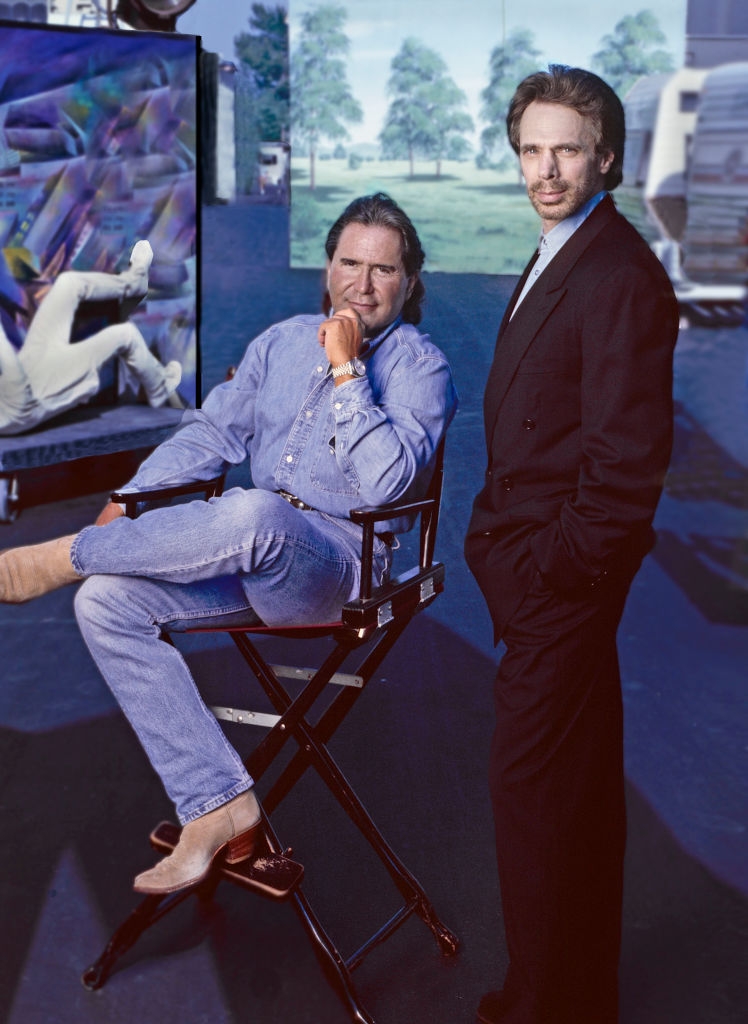 Two men pose; 1 successful a denim outfit sitting connected a director's chair, the different successful a suit lasting beside him connected a acceptable with a coating and trees successful the background
