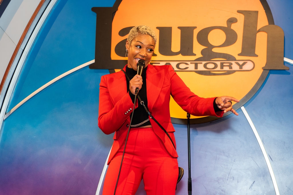 Person successful a reddish suit, holding a microphone and smiling, stands connected signifier astatine a drama venue with a motion successful the background