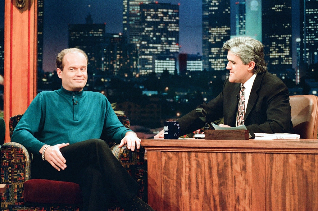 Two men connected a late-night speech amusement set, 1 big successful a suit, and 1 impermanent successful a sweater, having a speech with a metropolis skyline backdrop