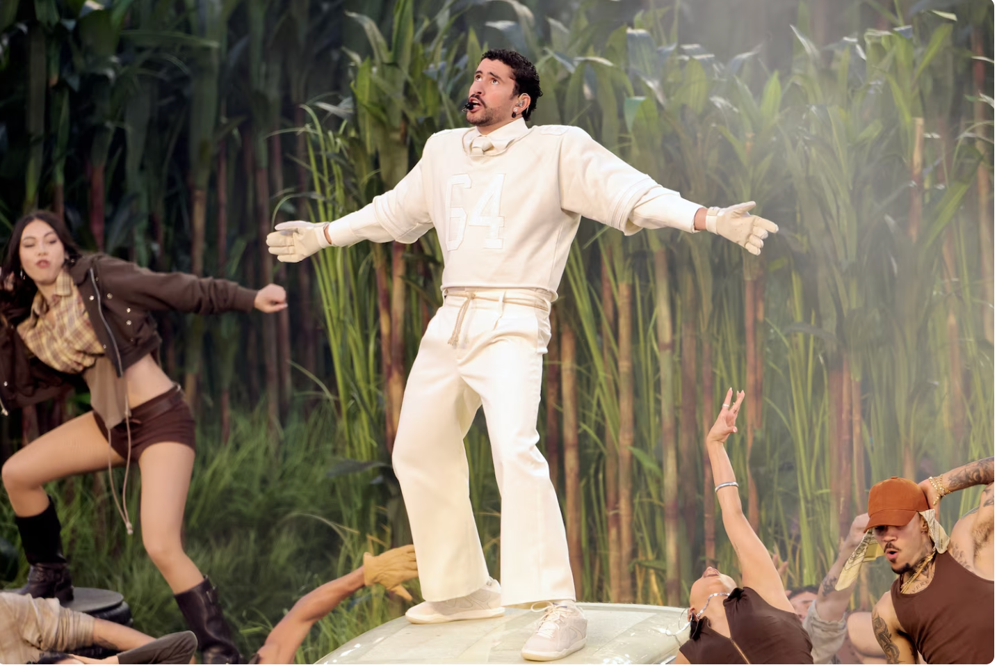 Artista enérgico actuando sobre un escenario rodeado de bailarines, con un atuendo blanco y guantes, en un ambiente de naturaleza