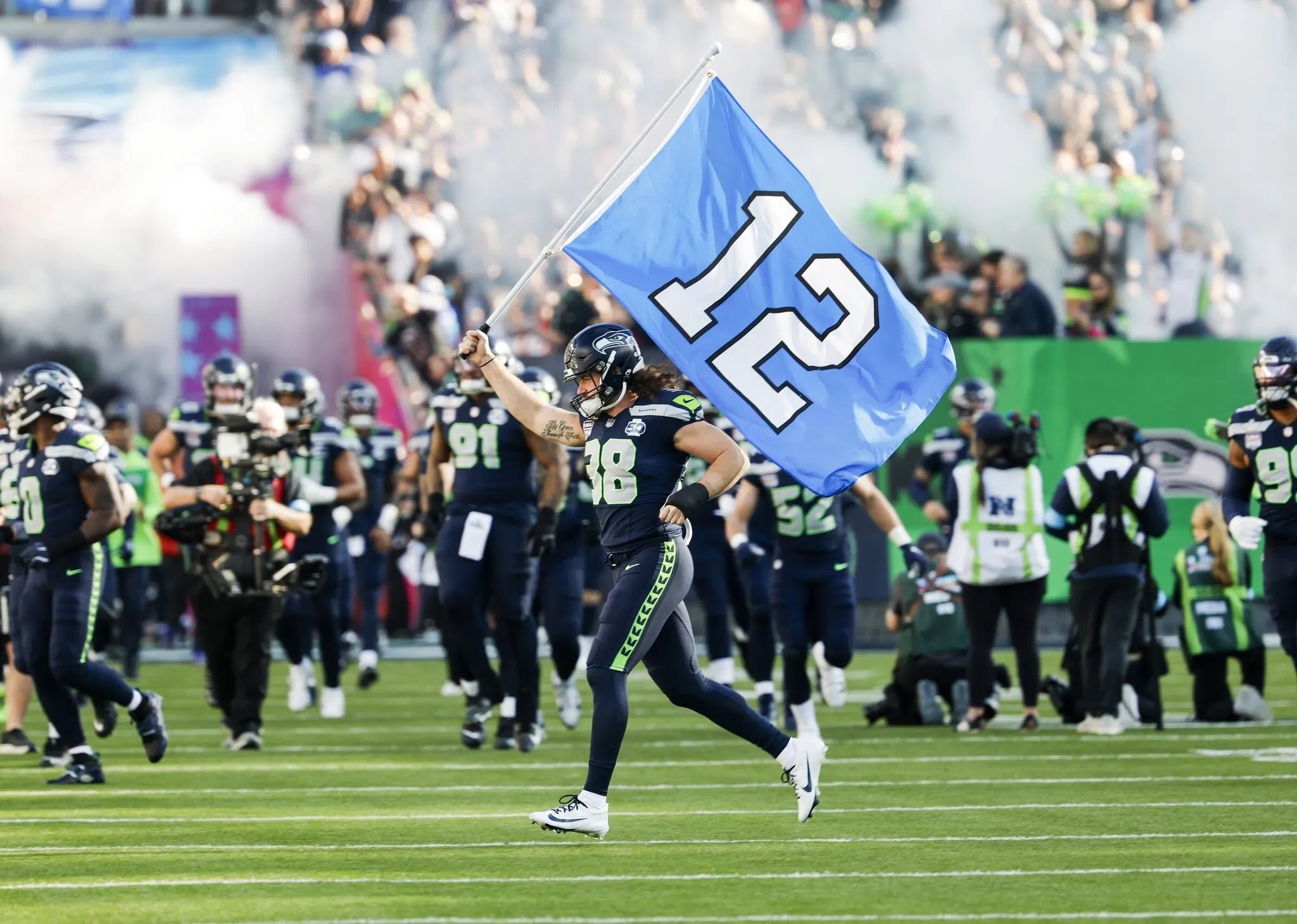 Jugador de fútbol americano del equipo Seahawks corre en el campo sosteniendo una bandera con el número 12, rodeado por compañeros y público