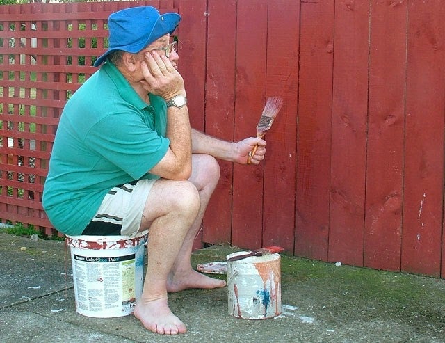 Persona sentada en un bote de pintura, sosteniendo un pincel, mirando una cerca sin pintar. Lleva gorra y camiseta