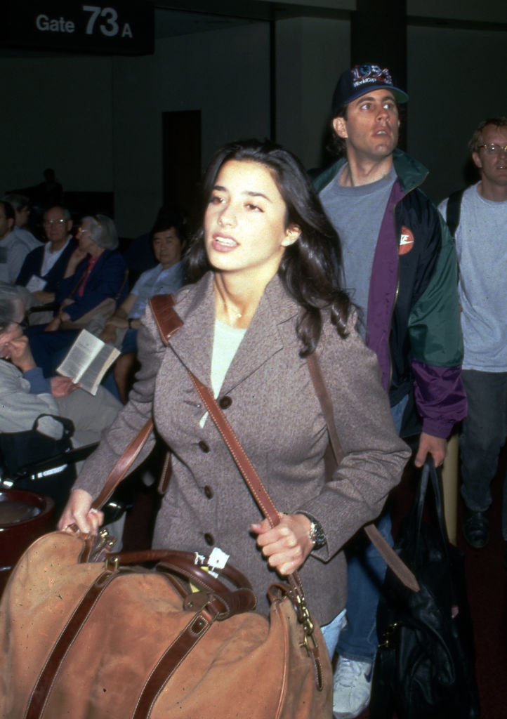 Woman with agelong hairsbreadth successful a blazer, carrying a ample container astatine an airdrome gate, with radical seated successful the background