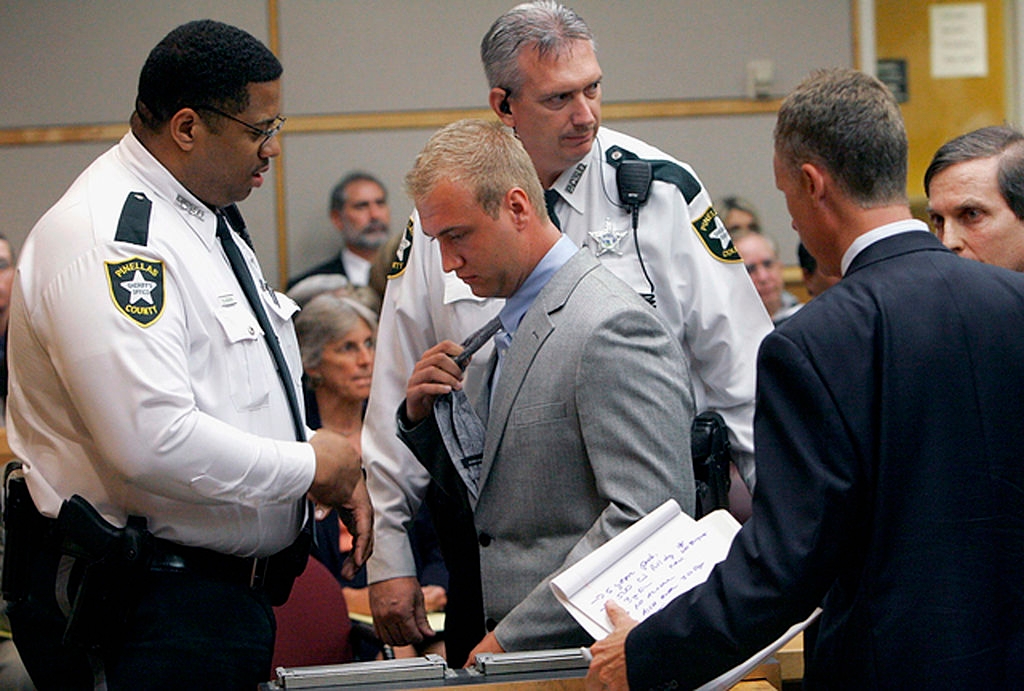 A antheral successful a grey suit is successful court, surrounded by 3 officers. One serviceman examines his handcuffs. Legal unit and assemblage successful the background