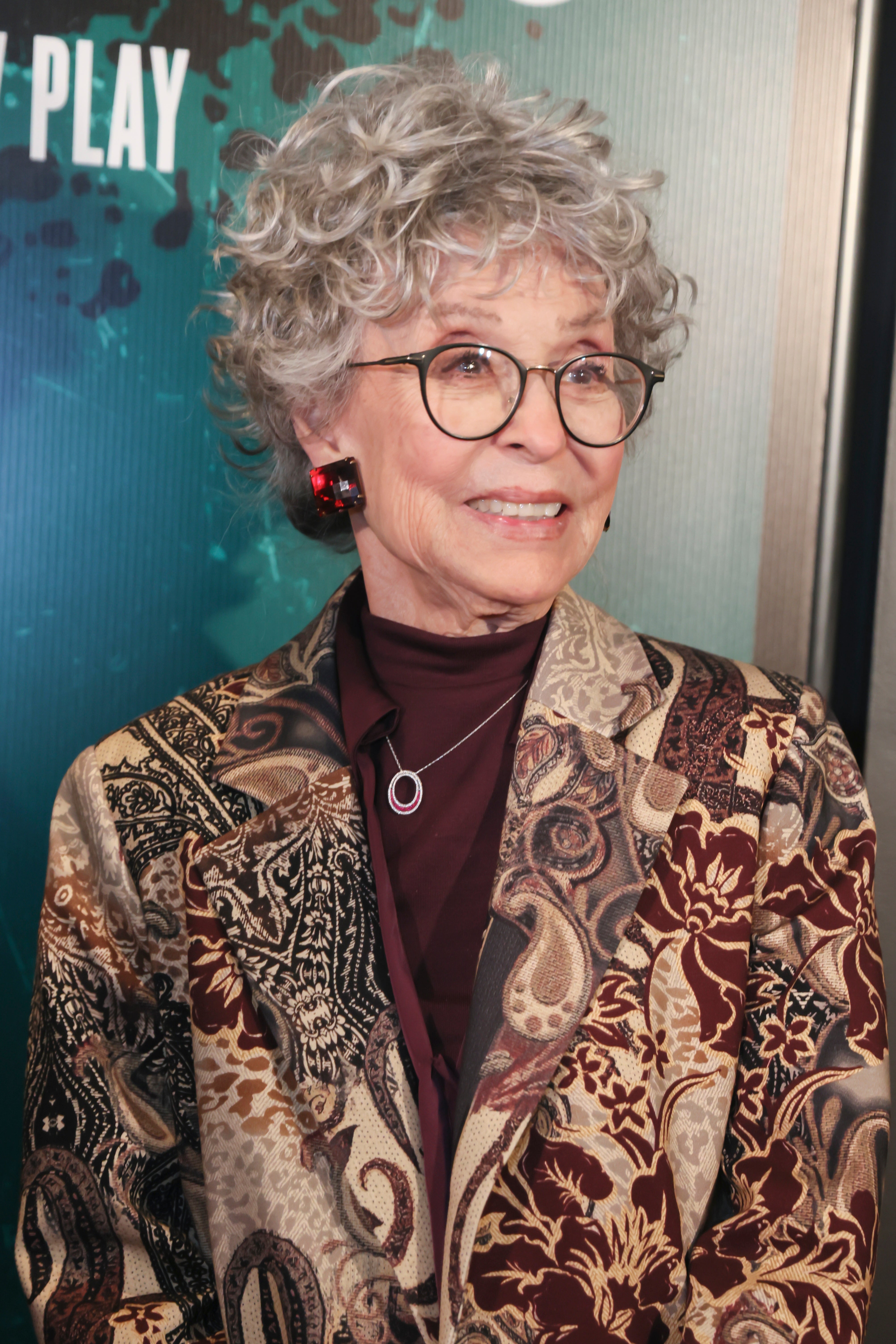 Person with curly hair, glasses, and a patterned blazer attends an event. They are smiling slightly, facing slightly to the left