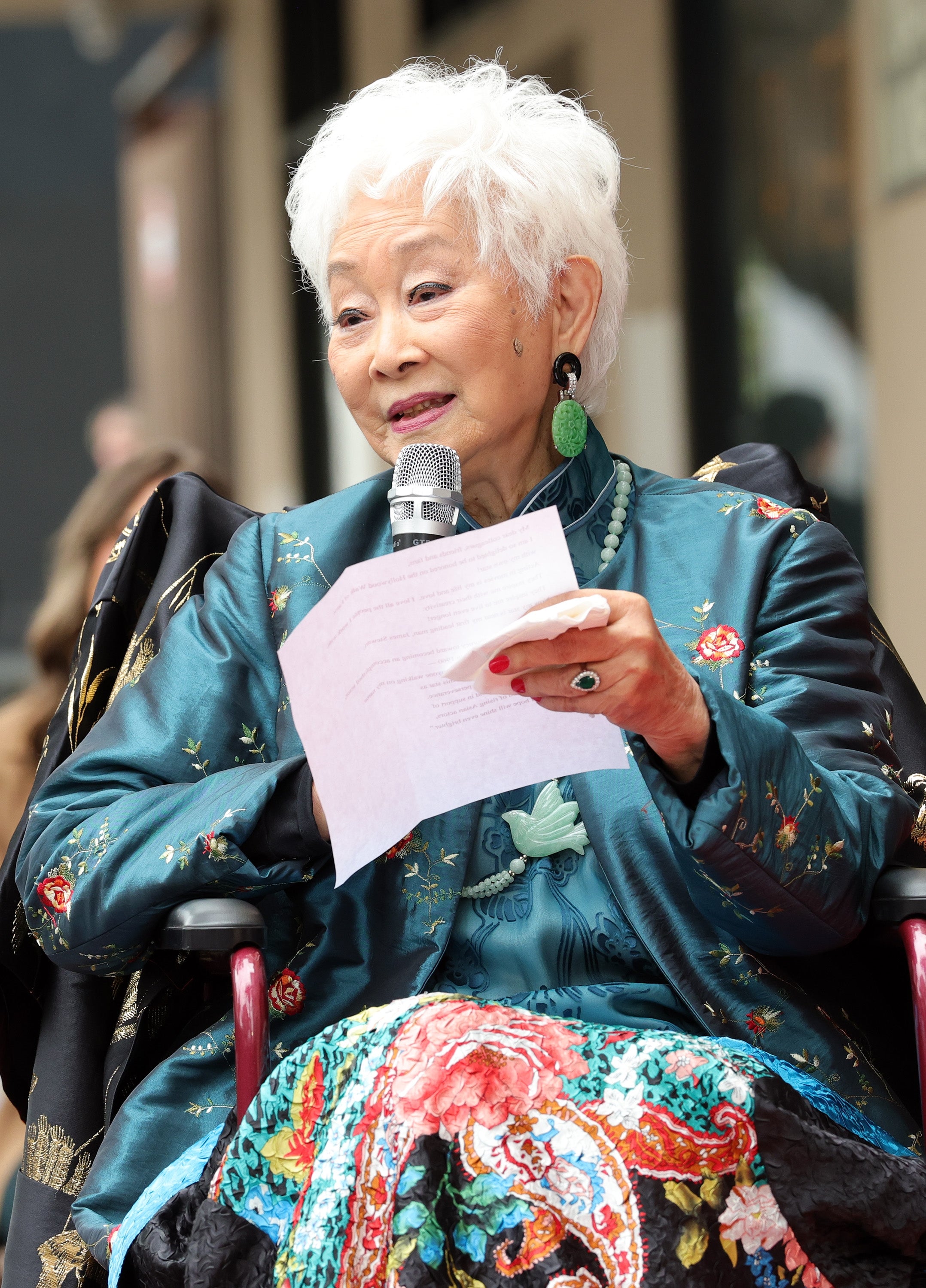Elderly person in a stylish embroidered outfit holds a microphone, speaking while seated and holding a piece of paper