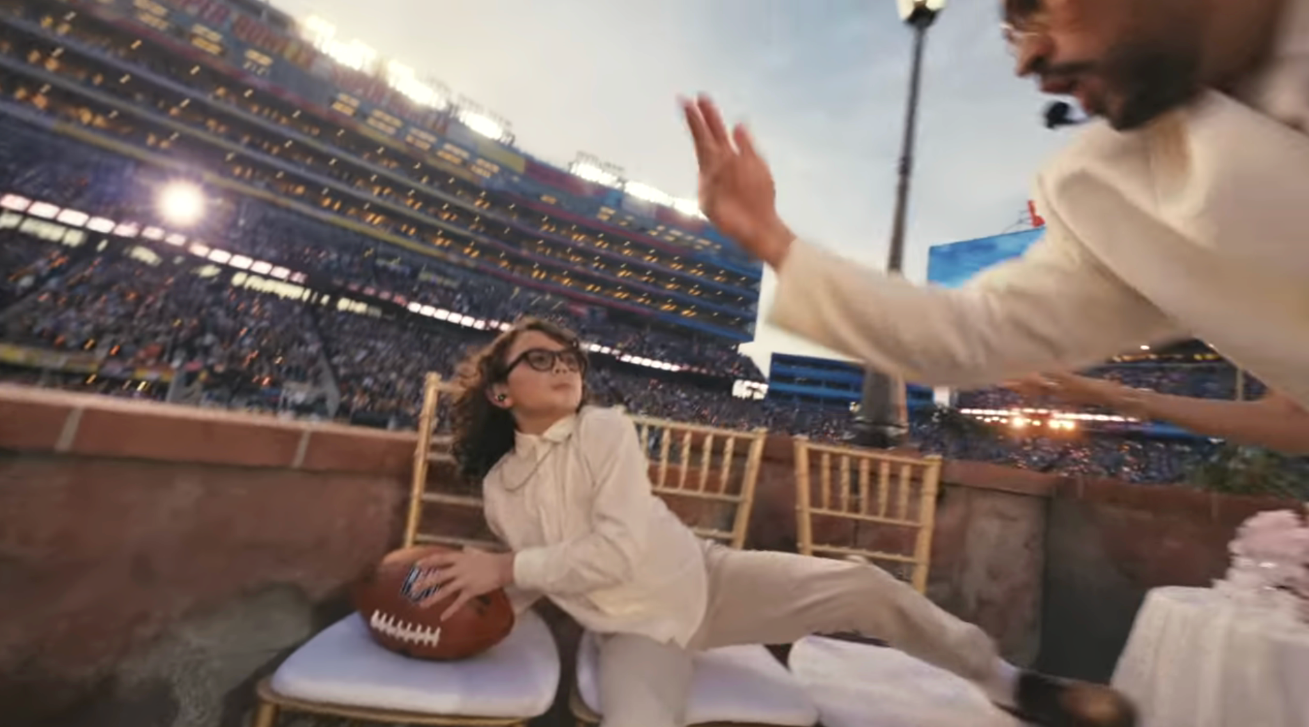 A kid  successful  a suit   and glasses sits by a shot   successful  a stadium suite, portion    an big  extends a manus  towards them