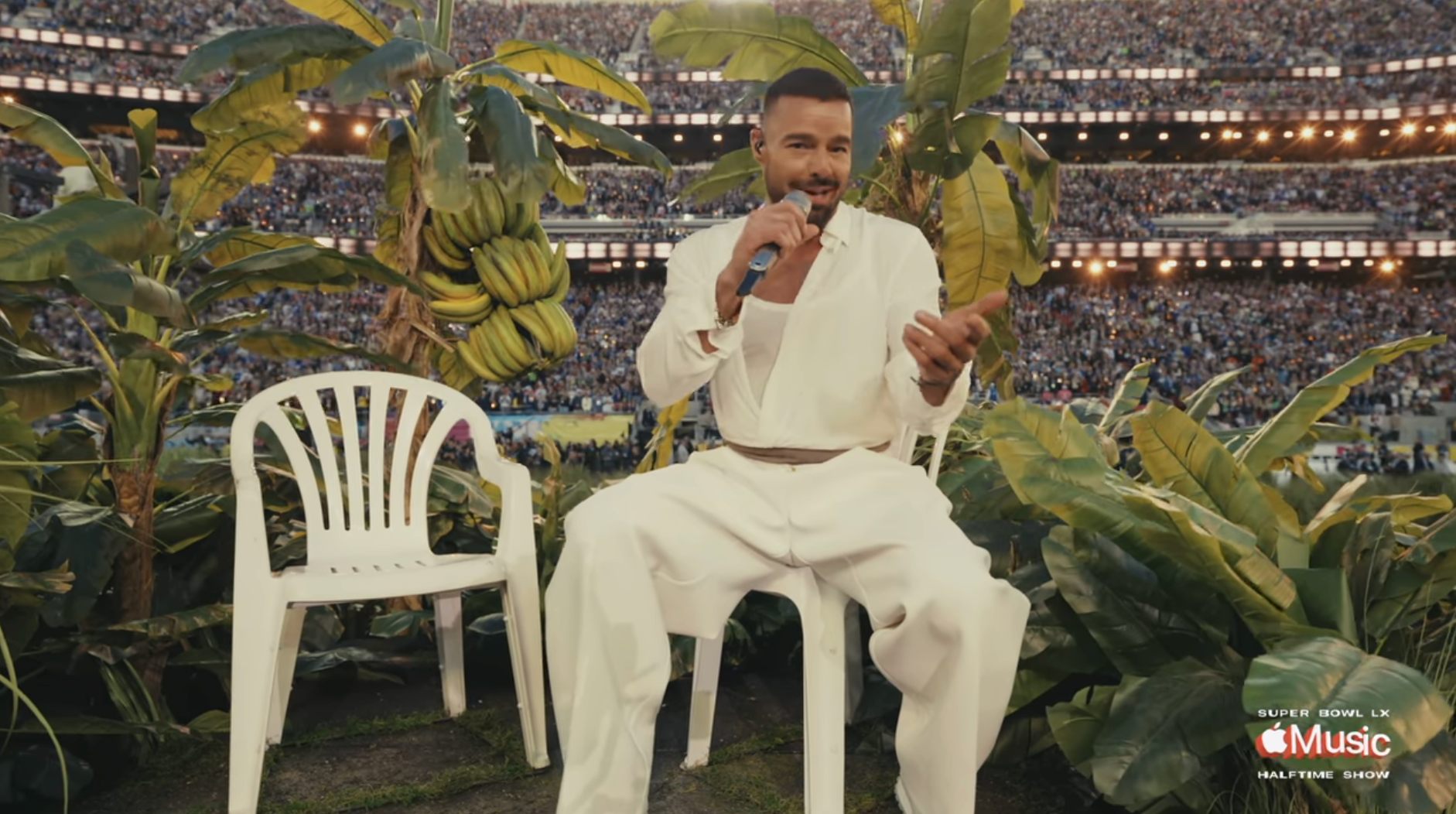 Person performs successful  a stadium, sitting among tropical plants connected  a achromatic  chair, wearing a achromatic  outfit, microphone successful  hand