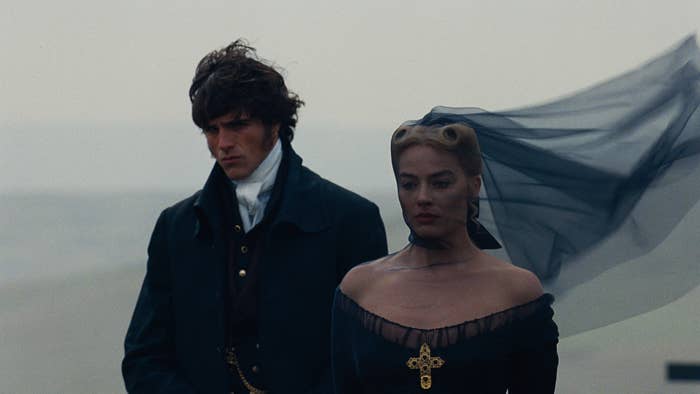 Two people in period attire stand side by side; the woman wears a dark off-shoulder dress with a veil